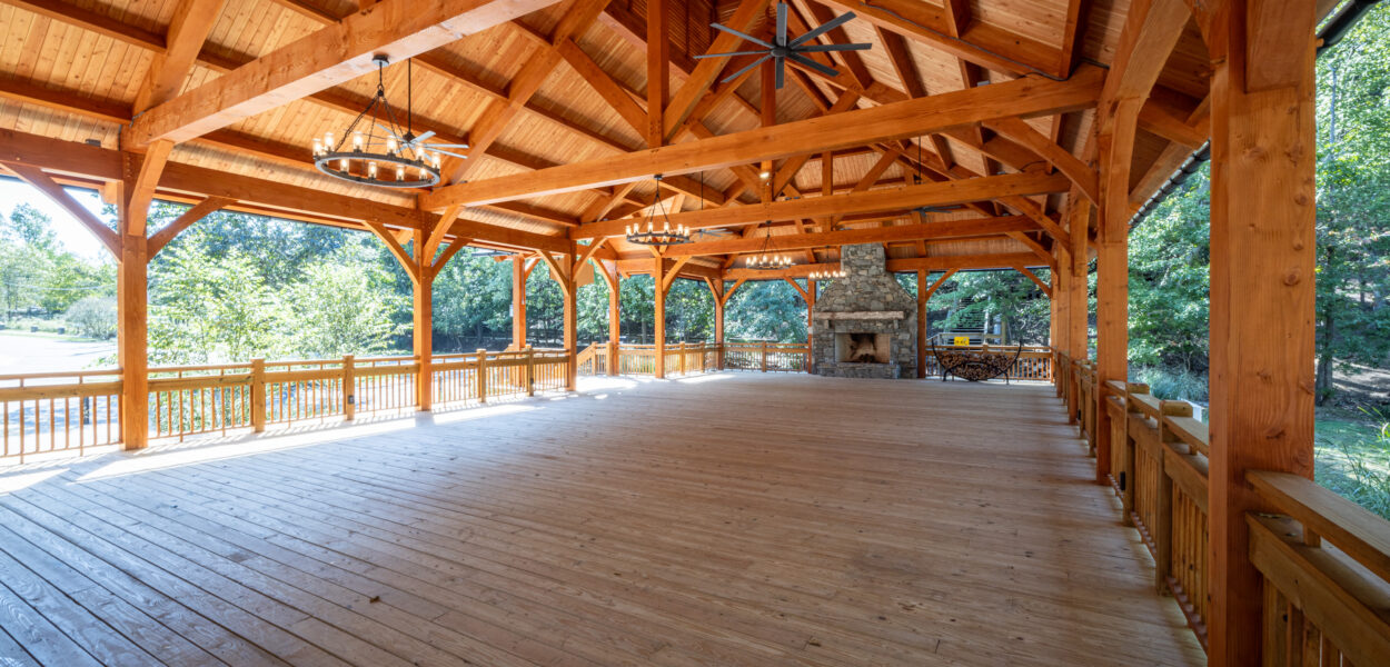 Spacious open-air wooden pavilion with a high timber roof, exposed beams, ceiling fans, and a stone fireplace at the far end, surrounded by a wooden railing, overlooking green trees.