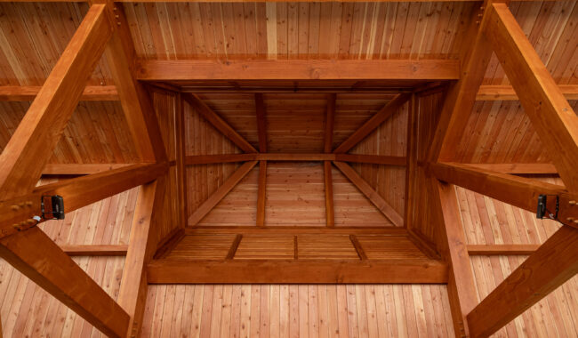 View looking up at a wooden ceiling with exposed beams and a rectangular recessed section in the center, showcasing the natural grain and warm tones of the wood.