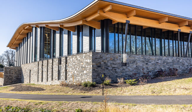 A modern building with a wavy wooden roof, large glass windows, and stone walls sits in a landscaped area with grass, shrubs, and a paved path under a clear blue sky.