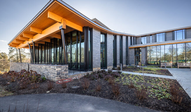 A modern building with large glass windows, wooden beams, and a landscaped garden in front. The structure features a slanted roof and a mix of stone and metal exterior, set against a clear sky.