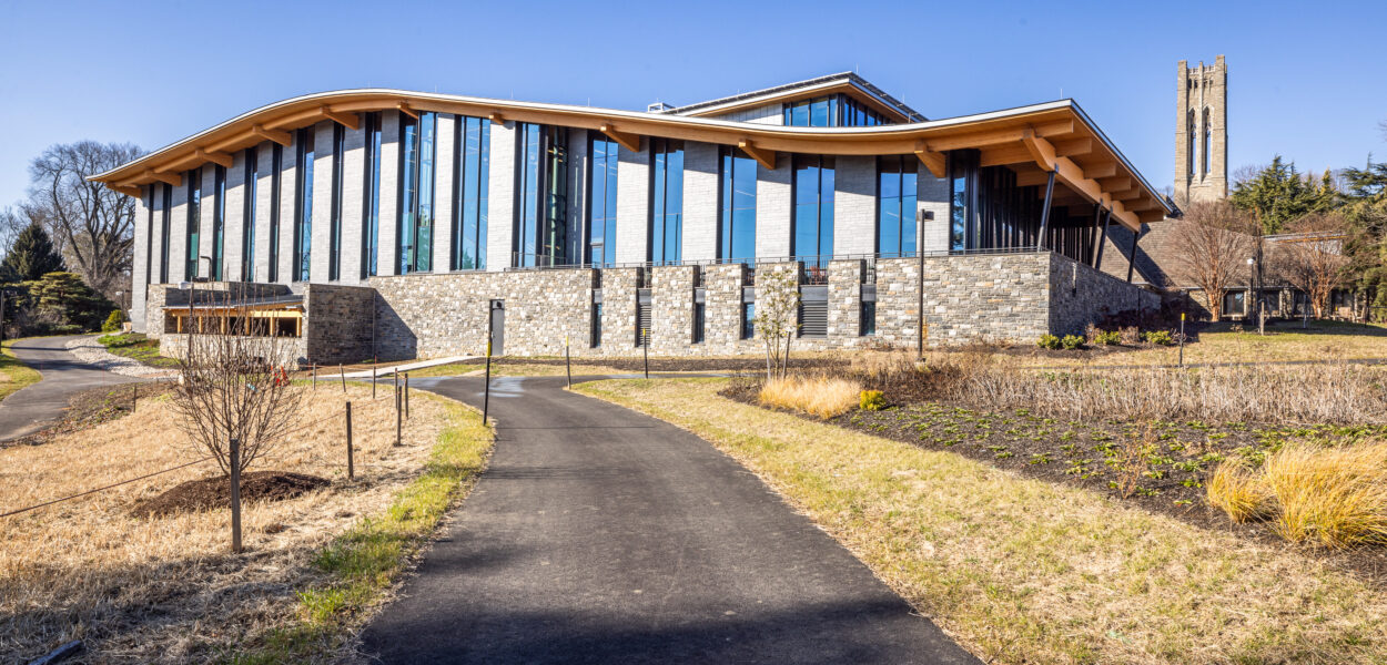 A modern building with large glass windows and a stone base sits beside a curving path, surrounded by landscaped grass and shrubs. A tall bell tower is visible in the background under a clear blue sky.