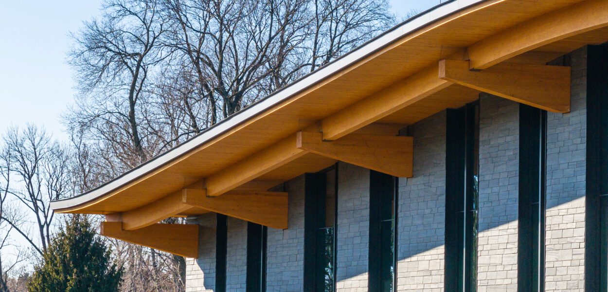 A modern building with a gently curved wooden roof, exposed beams, and tall vertical windows. Bare trees and evergreen trees are visible in the background under a clear blue sky.