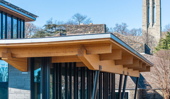 Modern building with a wooden overhang and angled metal supports, featuring large glass windows; stone buildings and a tall stone tower are visible in the background under a clear blue sky.
