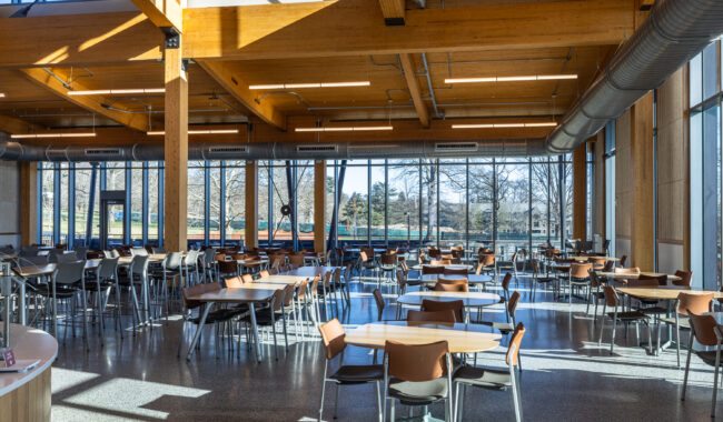 A spacious, sunlit cafeteria with wooden beams, large windows, and many empty tables and chairs arranged neatly throughout the room. Natural light fills the space, and trees are visible outside.