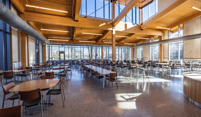 A spacious, modern cafeteria with wooden beams, large windows letting in natural light, and numerous empty tables and chairs arranged neatly throughout the room.