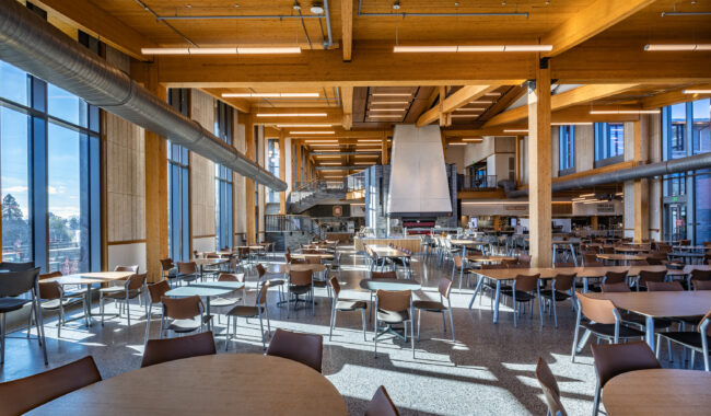 A spacious, modern cafeteria with wooden beams, floor-to-ceiling windows, and rows of empty tables and chairs. Sunlight fills the open area, highlighting the bright, airy atmosphere.