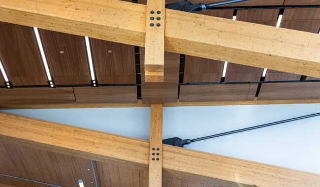 Wooden beams with visible bolts intersect on a modern ceiling, featuring wood panels, metal rods, and integrated linear lights. The design creates a geometric and architectural pattern overhead.