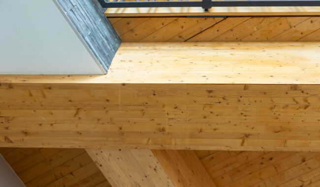 Large wooden beams intersect below an angled ceiling, with a metal railing and wooden floor above, showcasing exposed architecture and natural wood grain.