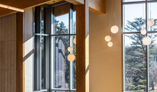 Large windows with wooden beams let in natural light, illuminating round pendant lights. Trees are visible outside through the glass, creating a bright, modern interior with a warm, natural feel.