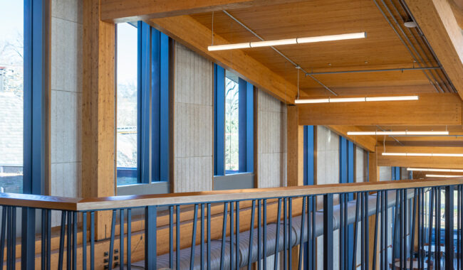 Modern interior with large vertical windows, wooden beams, and metal railing along an upper level. Overhead LED lights illuminate the space, highlighting the mix of wood and metal architectural elements.