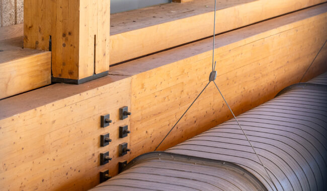 Close-up of wooden beams and posts joined with metal bolts, alongside a large curved metal duct suspended by cables, showcasing modern architectural and structural details.