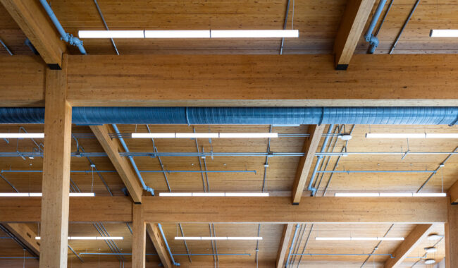 Exposed wooden ceiling beams with visible blue pipes and ductwork, illuminated by long, narrow ceiling lights in an industrial-style interior.