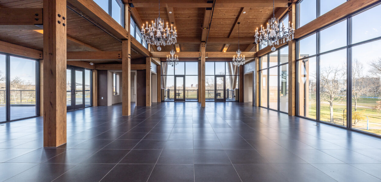 Spacious, empty hall with large windows, dark tiled floors, exposed wooden beams, and several elegant chandeliers hanging from the wooden ceiling. Sunlight streams in, illuminating the modern, open interior.