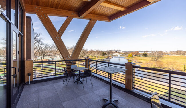 Modern balcony with wooden beams, metal railings, and outdoor tables and chairs overlooks a scenic park with a river and trees under a clear blue sky.