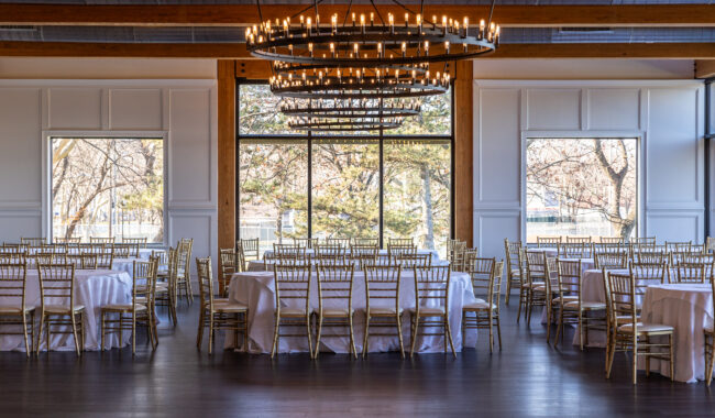 Elegant banquet hall set for an event, with round tables covered in white tablecloths and gold chairs, large windows letting in natural light, and two modern chandeliers hanging from a wooden ceiling.