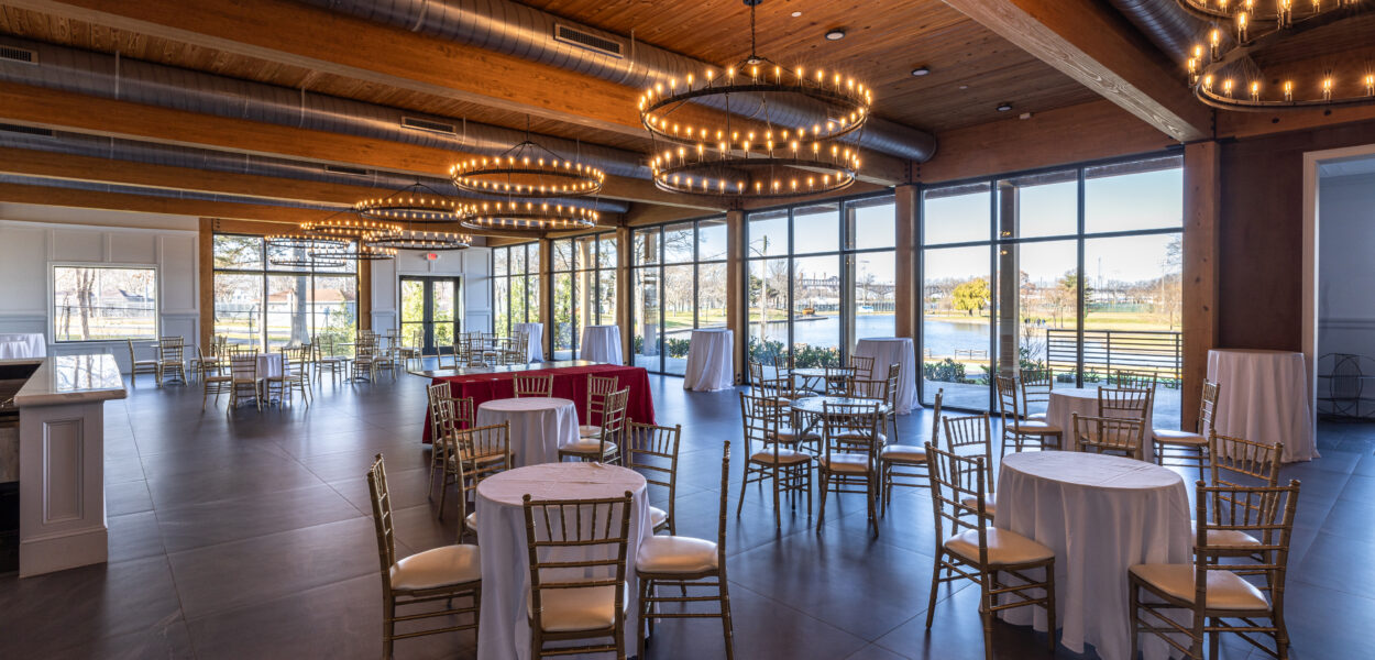 A spacious, modern event hall with large windows, round tables covered in white cloths, gold chairs, and ring-shaped chandeliers hanging from a wooden ceiling. Natural light fills the room.