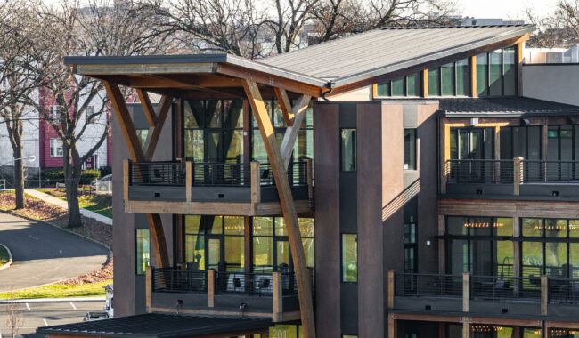A modern building featuring tall wooden support beams, large glass windows, and multiple balconies, with bare trees and a roadway in the background on a sunny day.