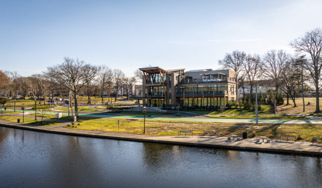 A modern, multi-story building with large glass windows stands beside a calm river, surrounded by grass, trees, benches, and walking paths on a sunny day.