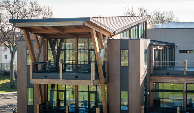 Modern building with large windows, wooden beams, and spacious balconies, featuring a unique angled roof. The structure is set against a backdrop of trees and a clear sky, with a parking lot visible below.