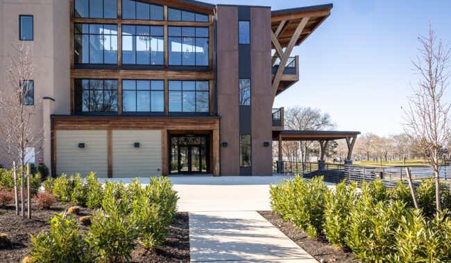 A modern, three-story building with large glass windows and wooden accents, surrounded by young landscaping and shrubs, with a paved walkway leading to the entrance on a sunny day.