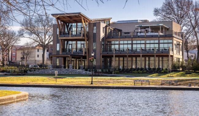 A modern, three-story building with large glass windows sits by a calm body of water, surrounded by grass, benches, trees, and a walking path under a clear sky.