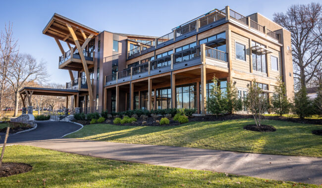 A modern, multi-story building with large windows, wooden and metal accents, and multiple balconies, surrounded by landscaped greenery and a driveway under a clear blue sky.