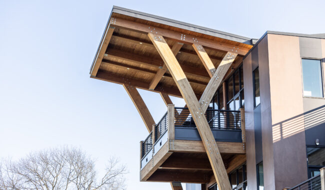 A modern building with large wooden beams supporting an overhanging roof and balcony, featuring metal railings and glass windows, set against a clear sky with bare tree branches visible.