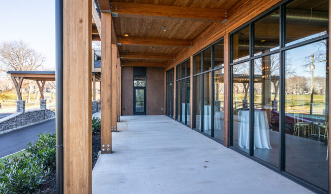 A modern building with large glass windows and wooden beams features a covered concrete patio. The area is bordered by plants and overlooks a grassy park with bare trees in the background.