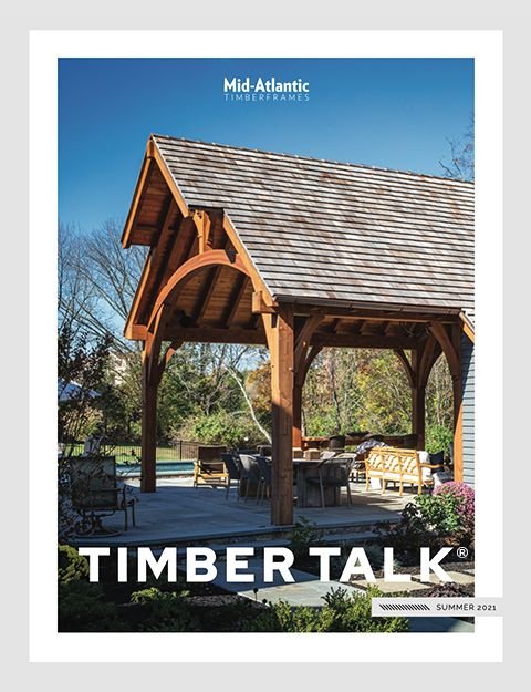 A wooden timber frame pavilion with outdoor seating beneath it, surrounded by green trees and shrubs under a clear blue sky. The cover reads Mid-Atlantic Timberframes, Timber Talk, and Summer 2021.